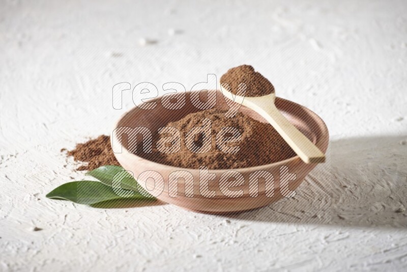 A wooden bowl and a wooden spoon full of cloves powder on a textured white flooring