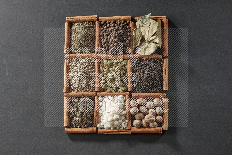 9 squares of cinnamon sticks full of cardamom in the middle surrounded by nutmeg, black pepper, bay laurel leaves, allspice, cumin, white pepper, dried basil and salt on black flooring
