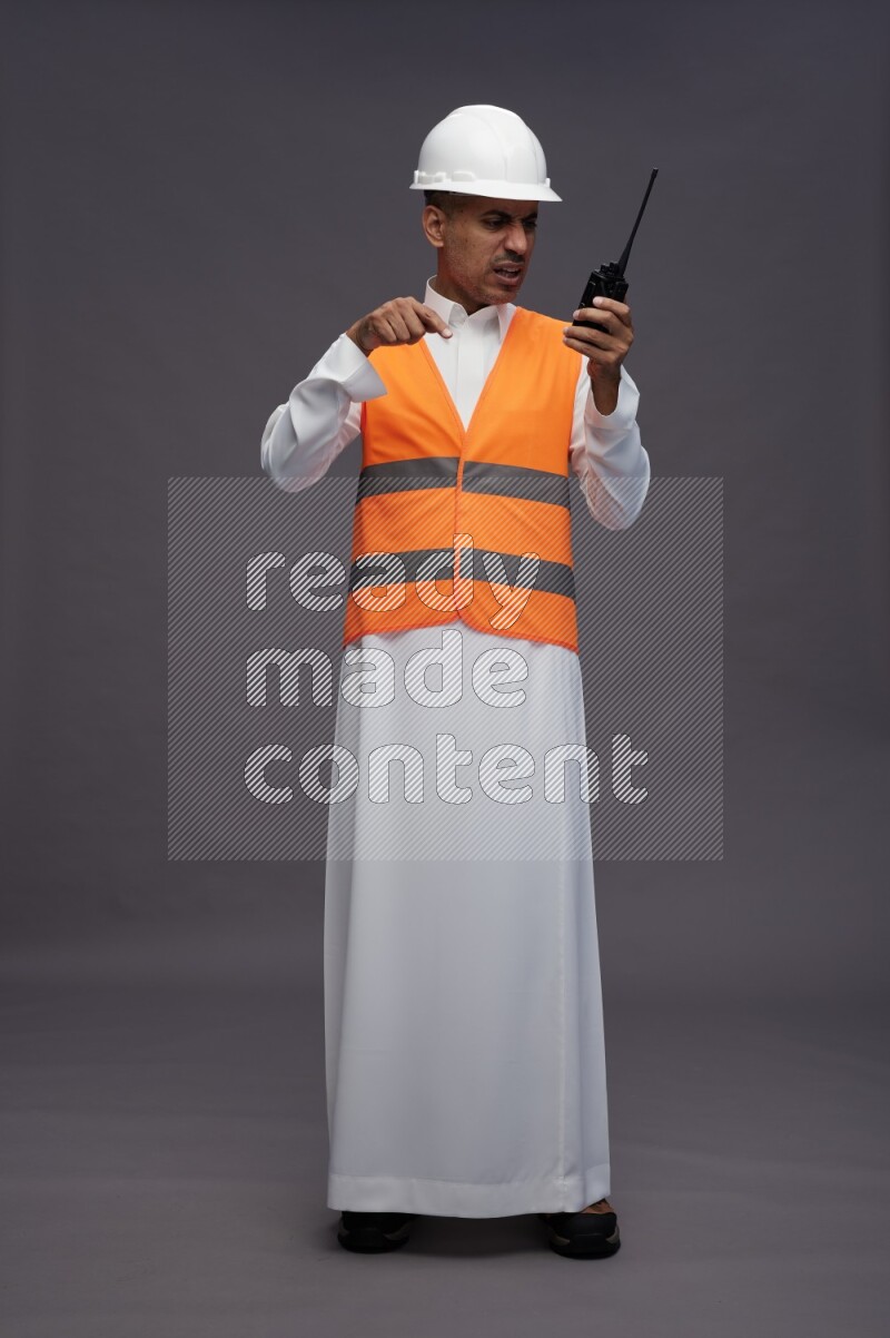 Saudi man wearing thob with engineer vest standing holding walkie-talkie on gray background
