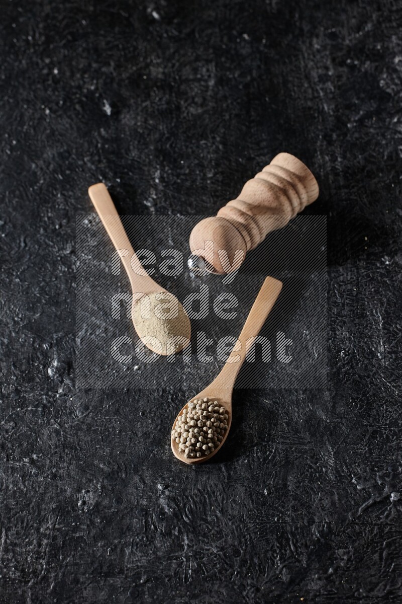 2 wooden spoons one full of white pepper powder and the other with pepper beads and a wooden pepper mill on textured black flooring