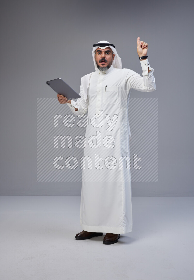 Saudi man Wearing Thob and white Shomag standing working on tablet on Gray background