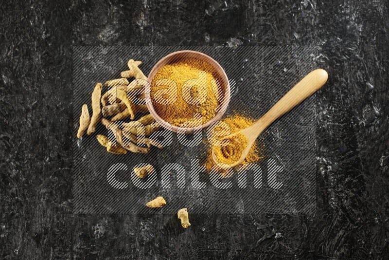 A wooden bowl and a wooden spoon full of turmeric powder with dried turmeric fingers on textured black flooring