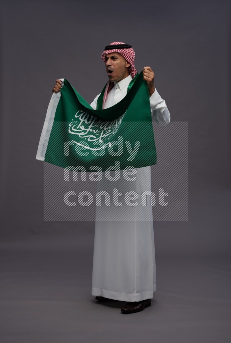 Saudi man wearing thob and shomag standing holding Saudi flag on gray background