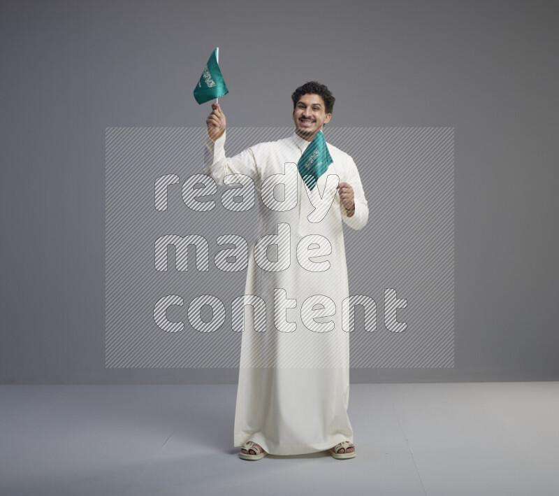 A Saudi man standing wearing thob holding small Saudi flag on gray background