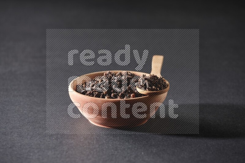 A wooden bowl and a wooden spoon full of cloves on a black flooring
