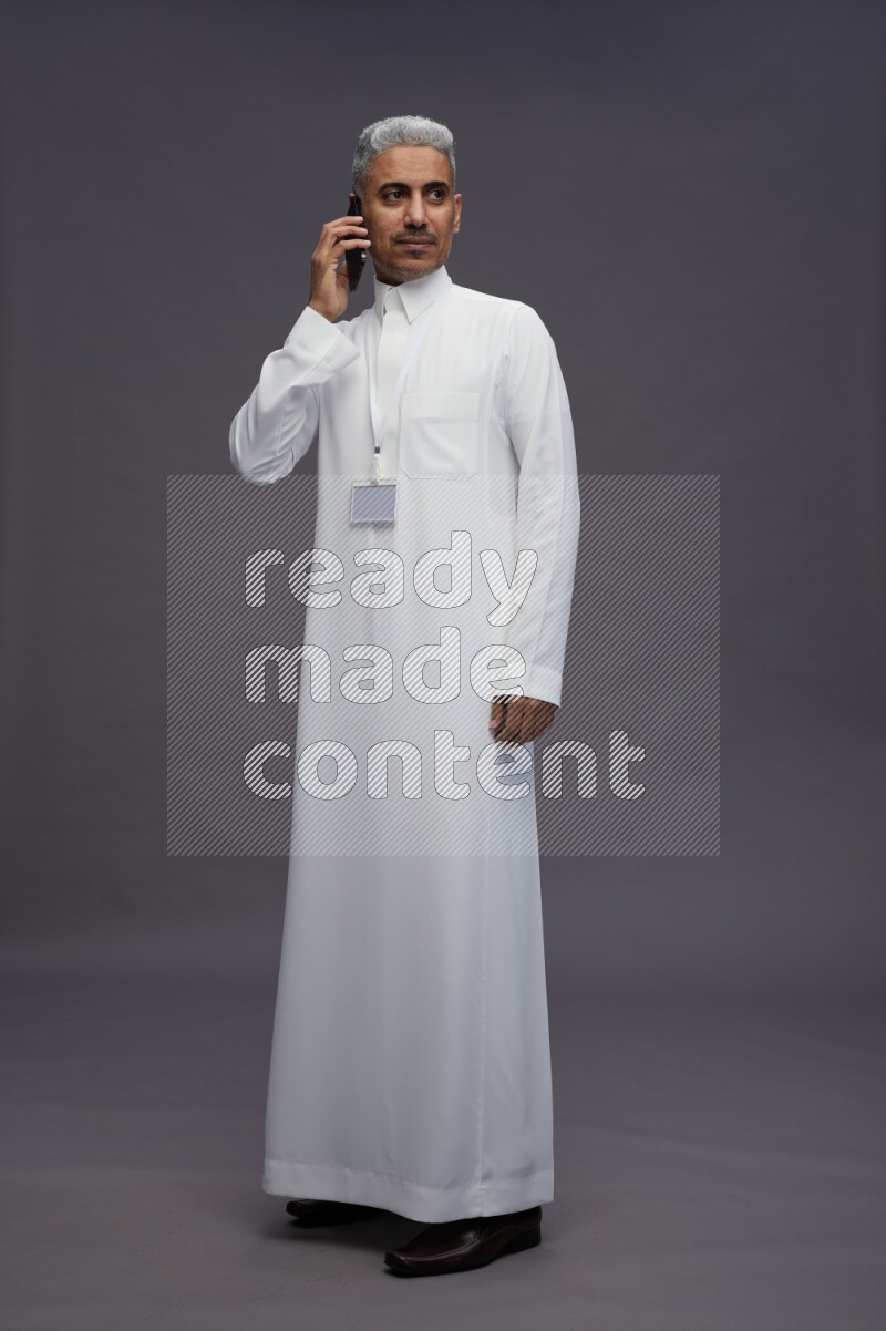 Saudi man wearing thob with neck strap employee badge standing talking on phone on gray background