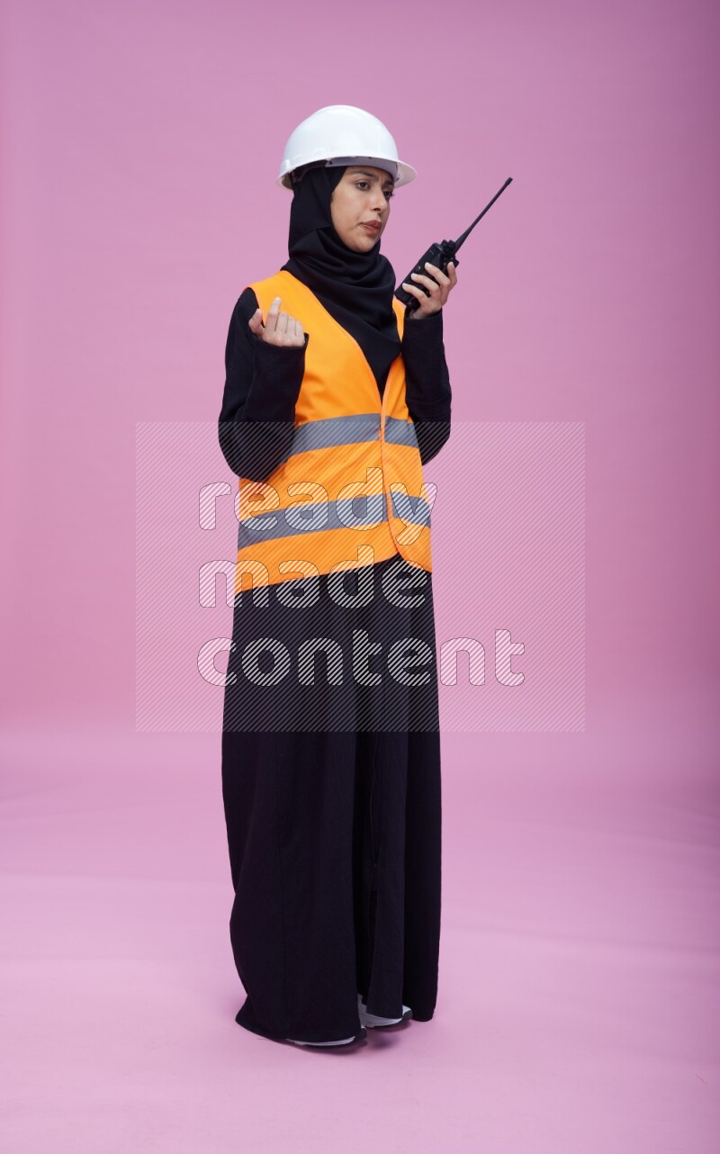 Saudi woman wearing Abaya with engineer vest and helmet standing holding walkie-talkie on pink background