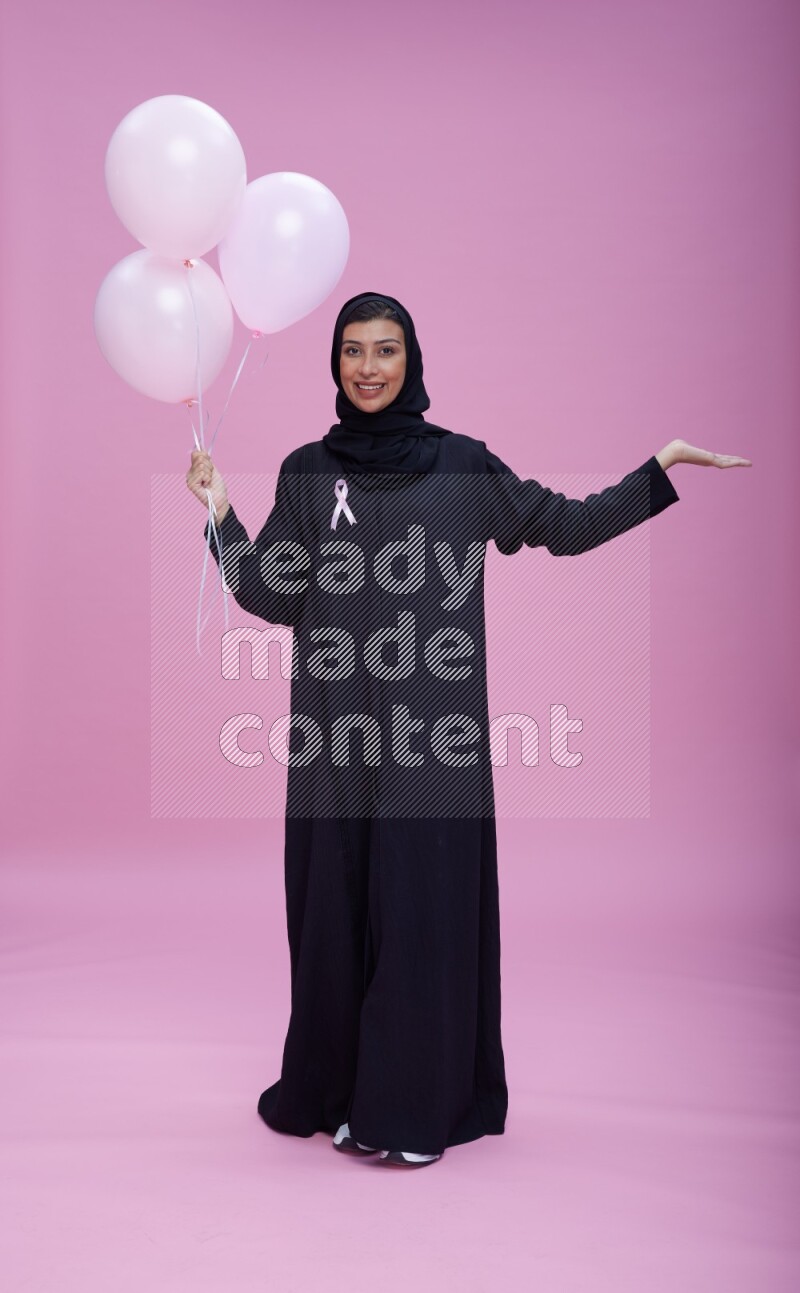 Saudi woman wearing pink ribbon on Abaya standing holding balloons on plain pink background