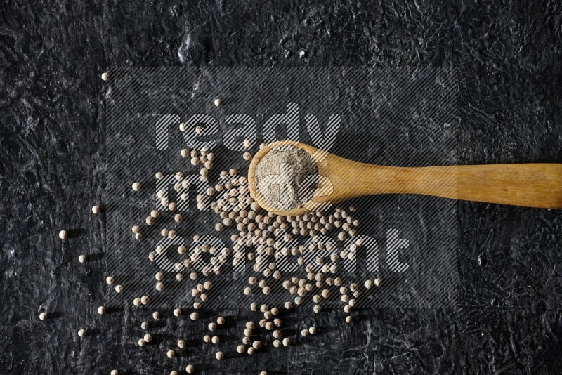 A wooden spoon full of white pepper powder with white pepper beads on textured black flooring