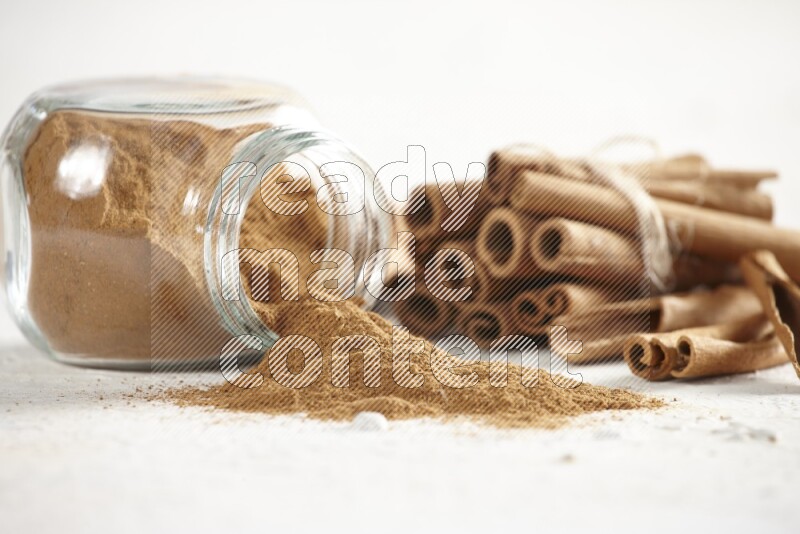 Flipped herbs glass jar full of cinnamon powder and cinnamon sticks in the back on a textured white background