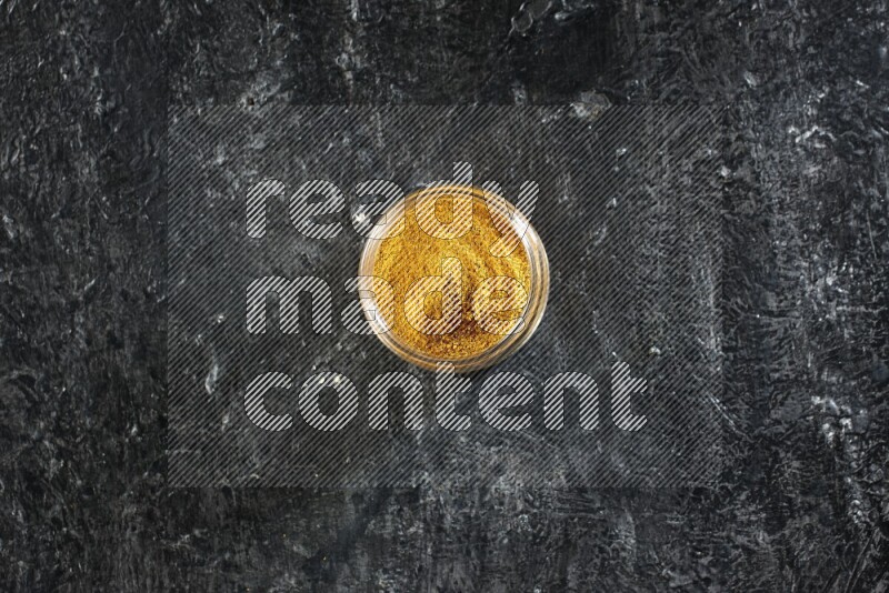 A glass jar full of turmeric powder on a textured black flooring