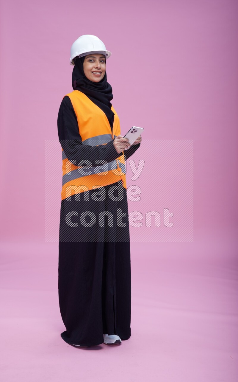 Saudi woman wearing Abaya with engineer vest and helmet standing texting on phone on pink background