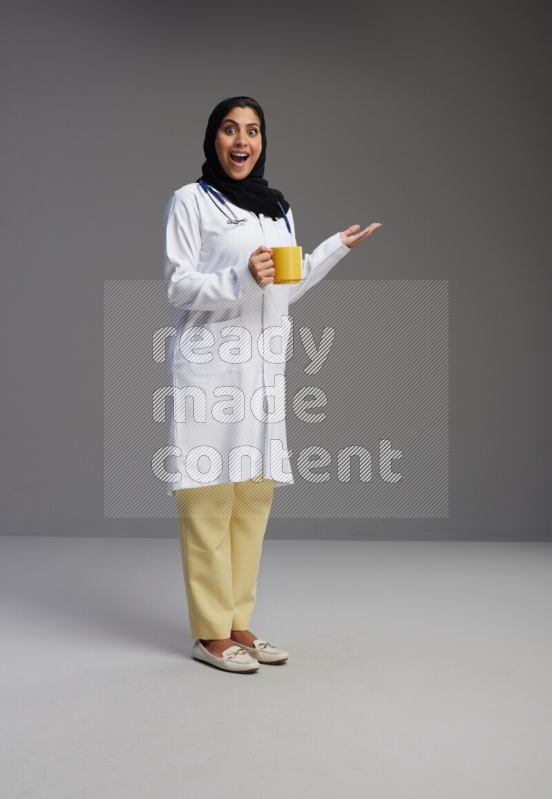 Saudi woman wearing lab coat with stethoscope standing holding mug on Gray background