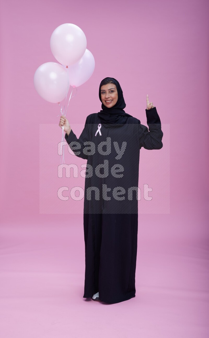 Saudi woman wearing pink ribbon on Abaya standing holding balloons on plain pink background