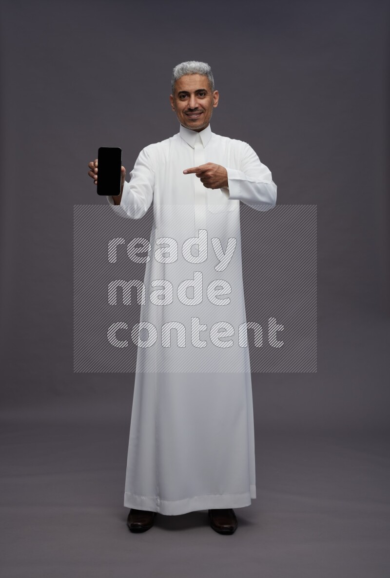 Saudi man wearing thob standing showing phone to camera on gray background