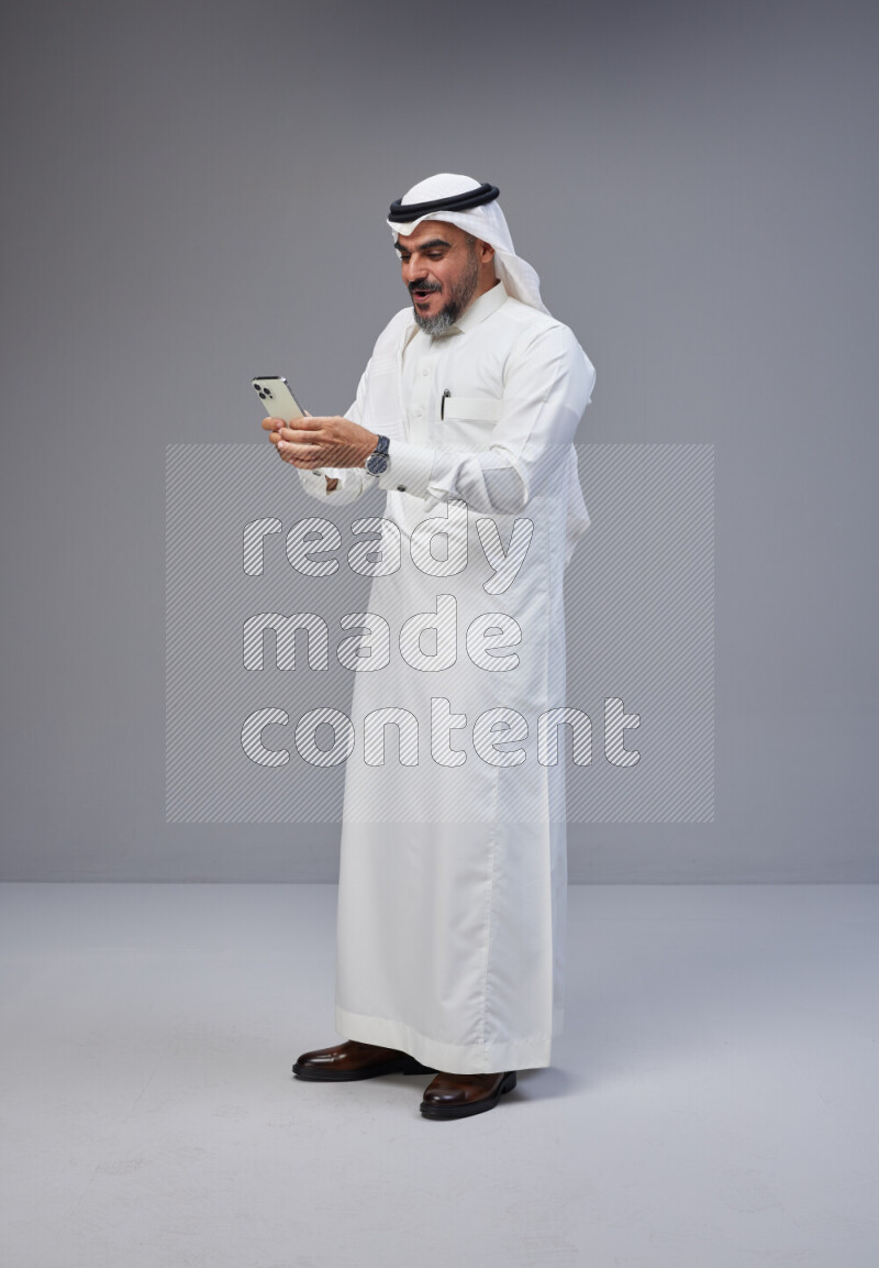 Saudi man Wearing Thob and white Shomag standing texting on phone on Gray background