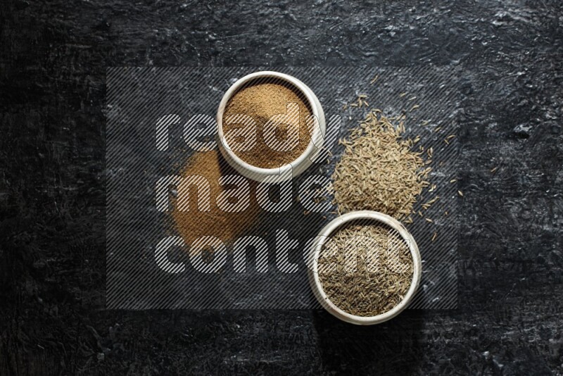 2 beige bowls, one full of cumin powder and the other full of seeds on a textured black flooring