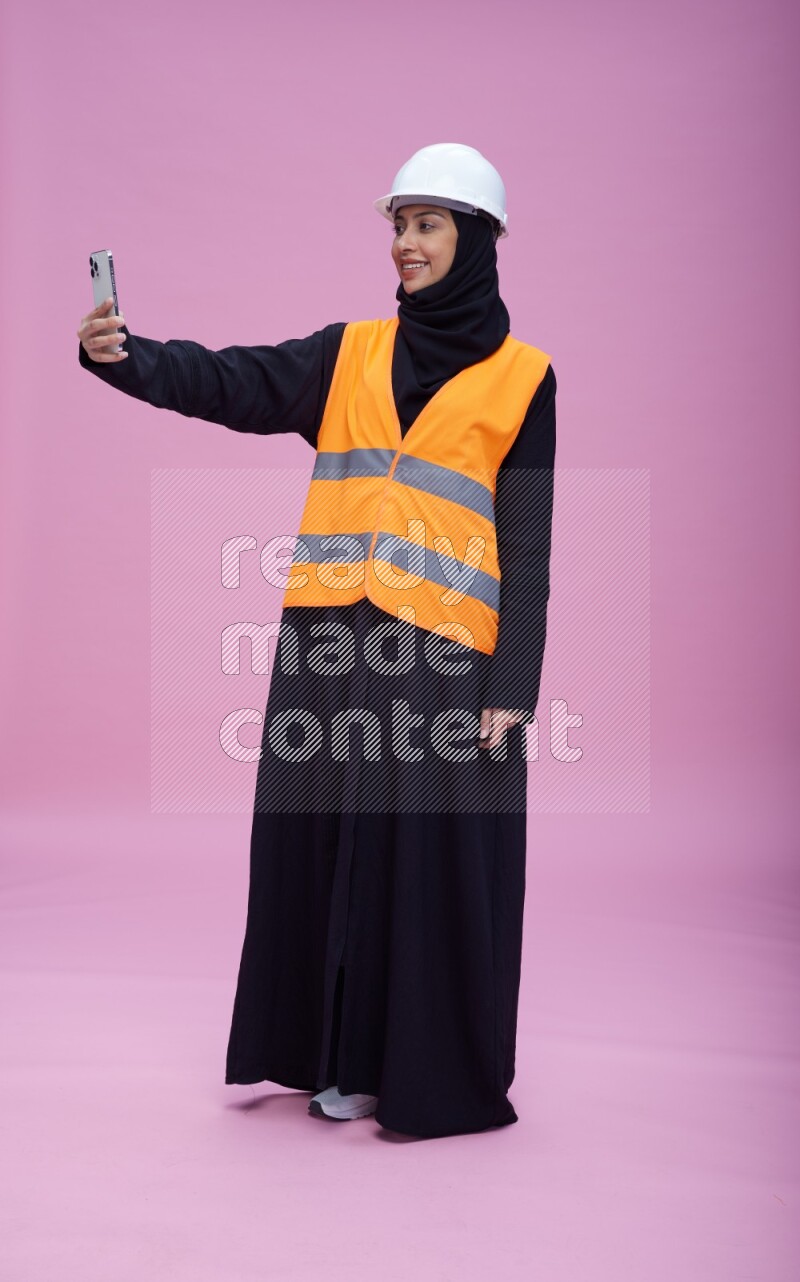 Saudi woman wearing Abaya with engineer vest and helmet standing taking selfie on pink background