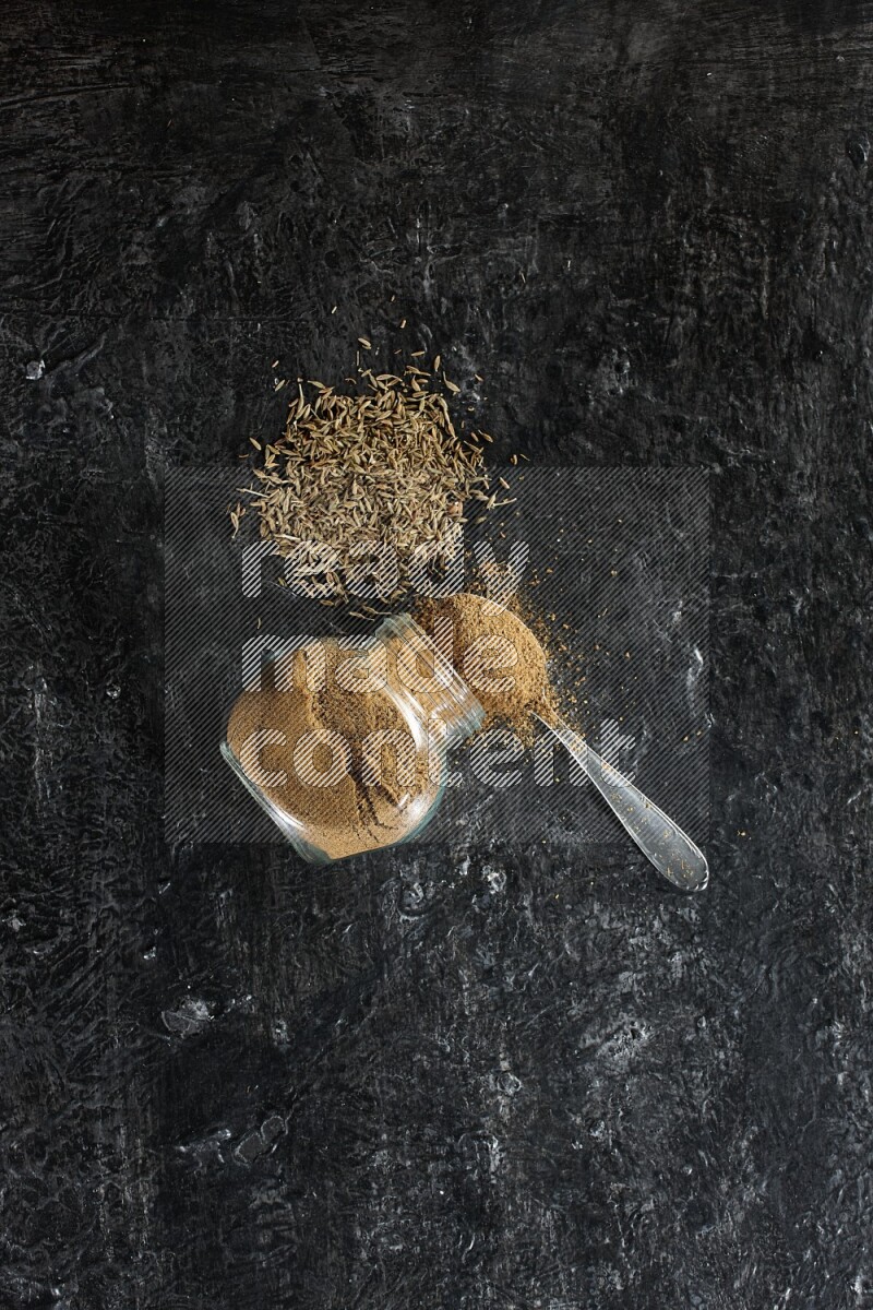A flipped glass spice jar and a metal spoon full of cumin powder and powder spilled out with cumin seeds on a textured black flooring