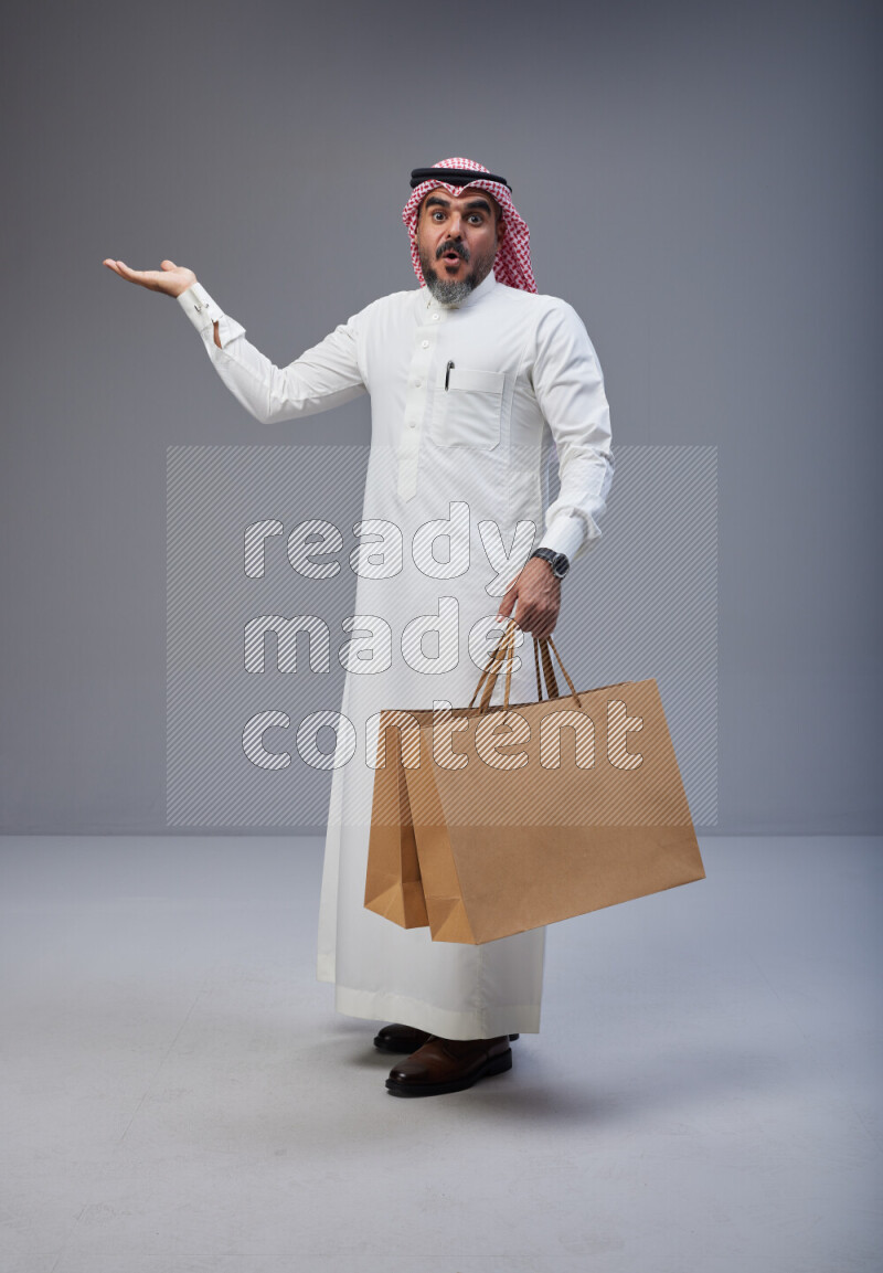 Saudi man Wearing Thob and red Shomag standing holding shopping bag on Gray background