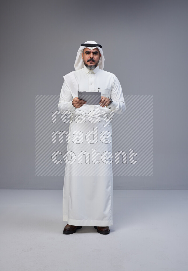 Saudi man Wearing Thob and white Shomag standing working on tablet on Gray background