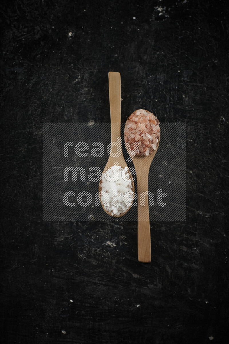2 wooden spoons filled with fine and coarse salt on black background