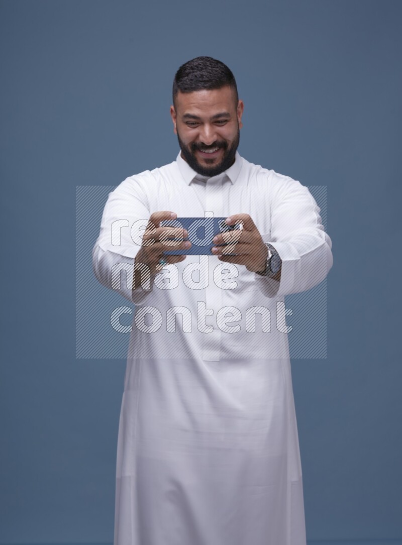 A man Playing a Game on his Smartphone  on Blue Background wearing Saudi Thob