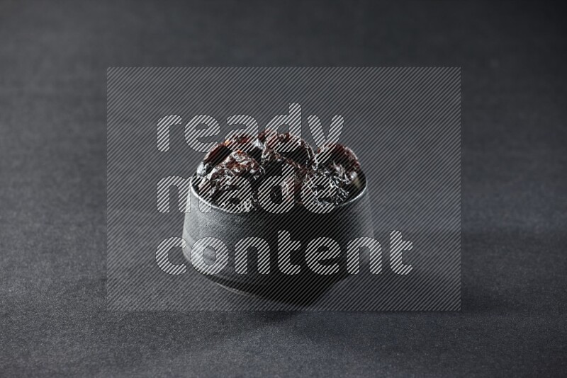 A black pottery bowl full of dried plums on a black background in different angles
