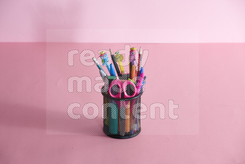 Pens, pencils and other different school supplies in a metal pen holder on rose background