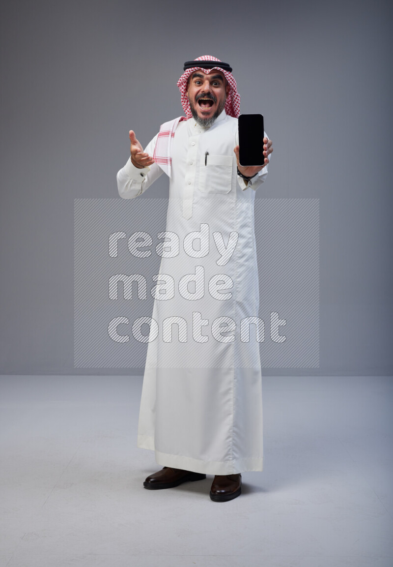 Saudi man Wearing Thob and red Shomag standing showing phone to camera on Gray background