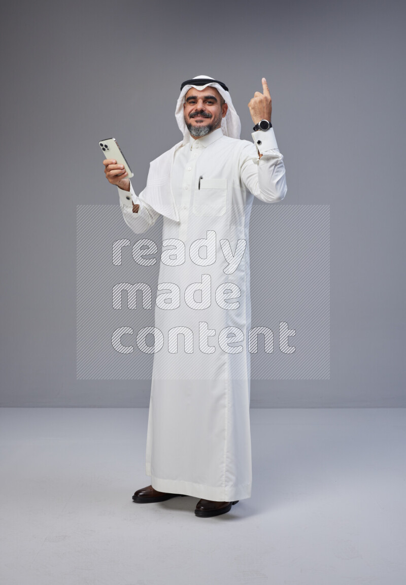 Saudi man Wearing Thob and white Shomag standing texting on phone on Gray background