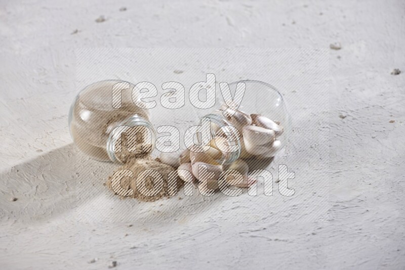 2 glass spice jars, one full of garlic powder and the other full of garlic cloves both flipped on a textured white flooring