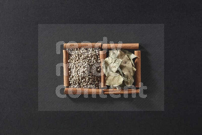 مربعان من أعواد القرفة ممتلئان بحبوب الفلفل الأبيض وأوراق الغار علي خلفية سوداء