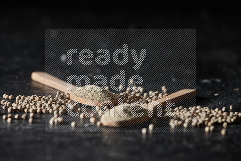 2 wooden spoons full of white pepper powder with white pepper beads on textured black flooring