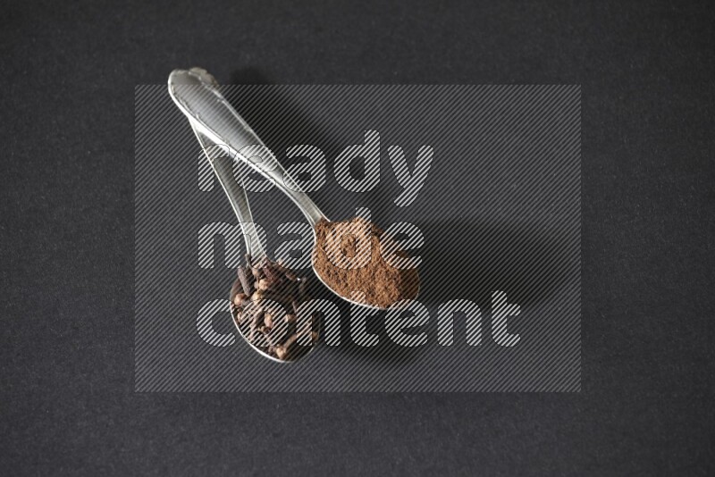 2 metal spoons, one full of cloves grains and the other full of cloves powder on a black flooring