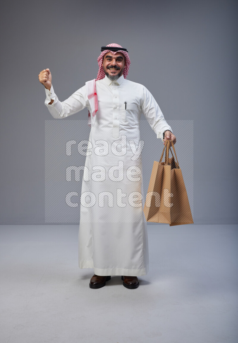 Saudi man Wearing Thob and red Shomag standing holding shopping bag on Gray background