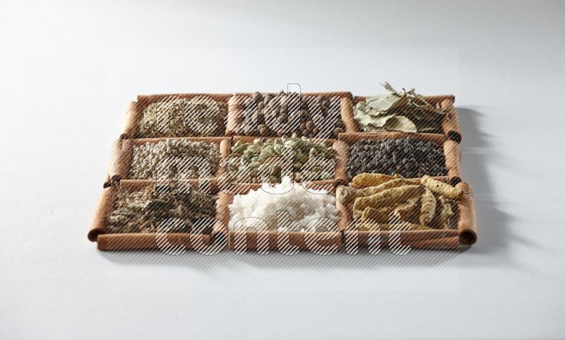 9 squares of cinnamon sticks full of cardamom in the middle surrounded by turmeric, salt, dried basil, white pepper, cumin, allspice, bay laurel leaves and black pepper on white flooring