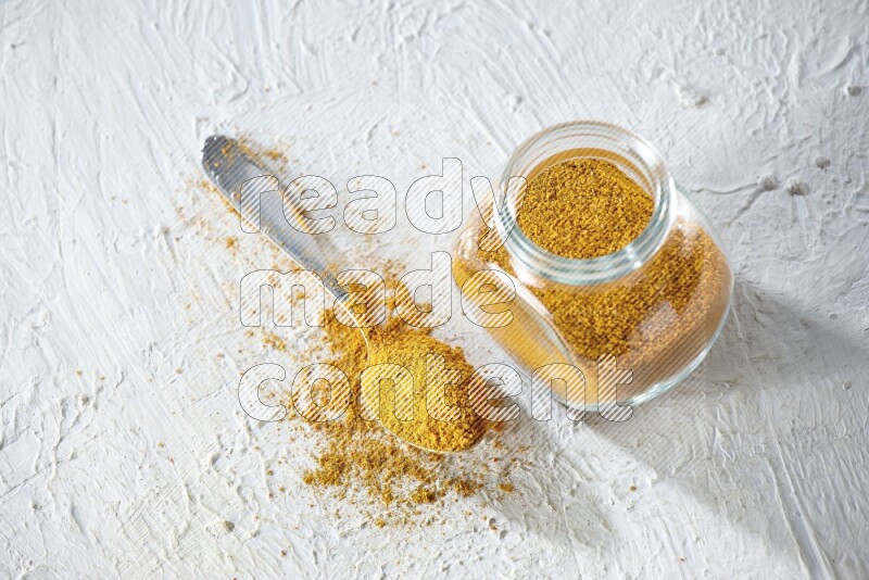A glass spice jar and metal spoon full of turmeric powder on textured white flooring