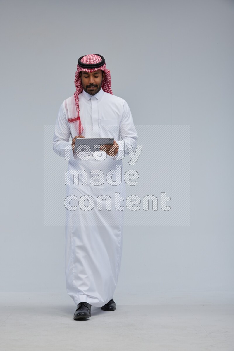 Saudi man Wearing Thob and shomag standing working on tablet on Gray background