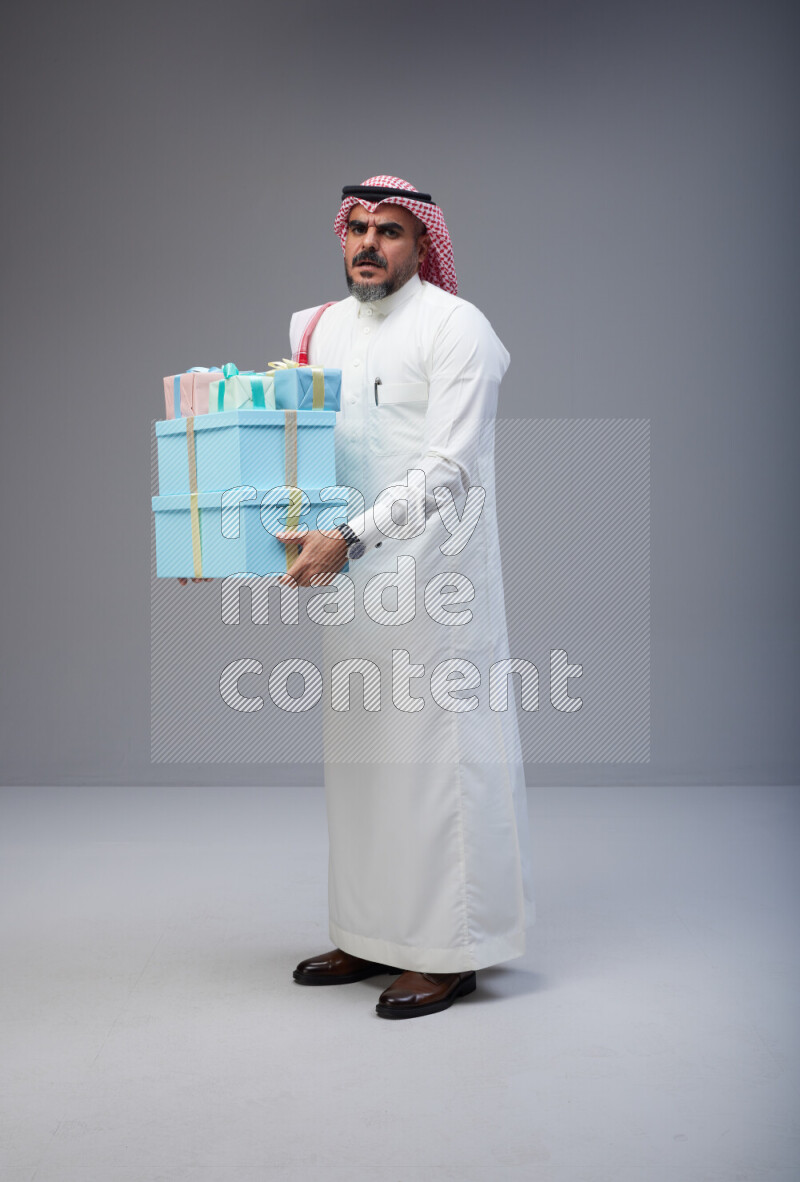 Saudi man Wearing Thob and red Shomag standing holding gift box on Gray background