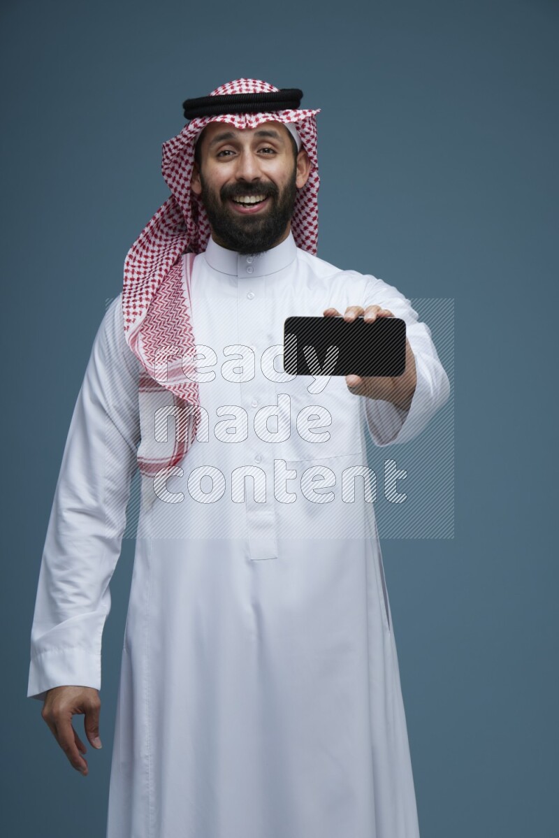 A man showing his phone Screen in a blue background wearing Saudi Thob with Shomag