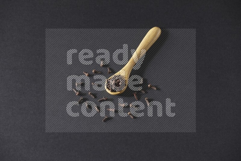 A wooden spoon full of cloves and some spreaded out of it on a black flooring