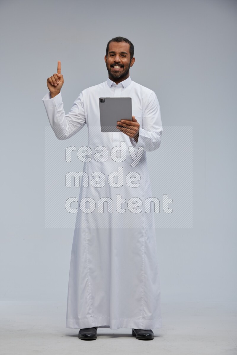 Saudi man wearing Thob standing working on tablet on Gray background