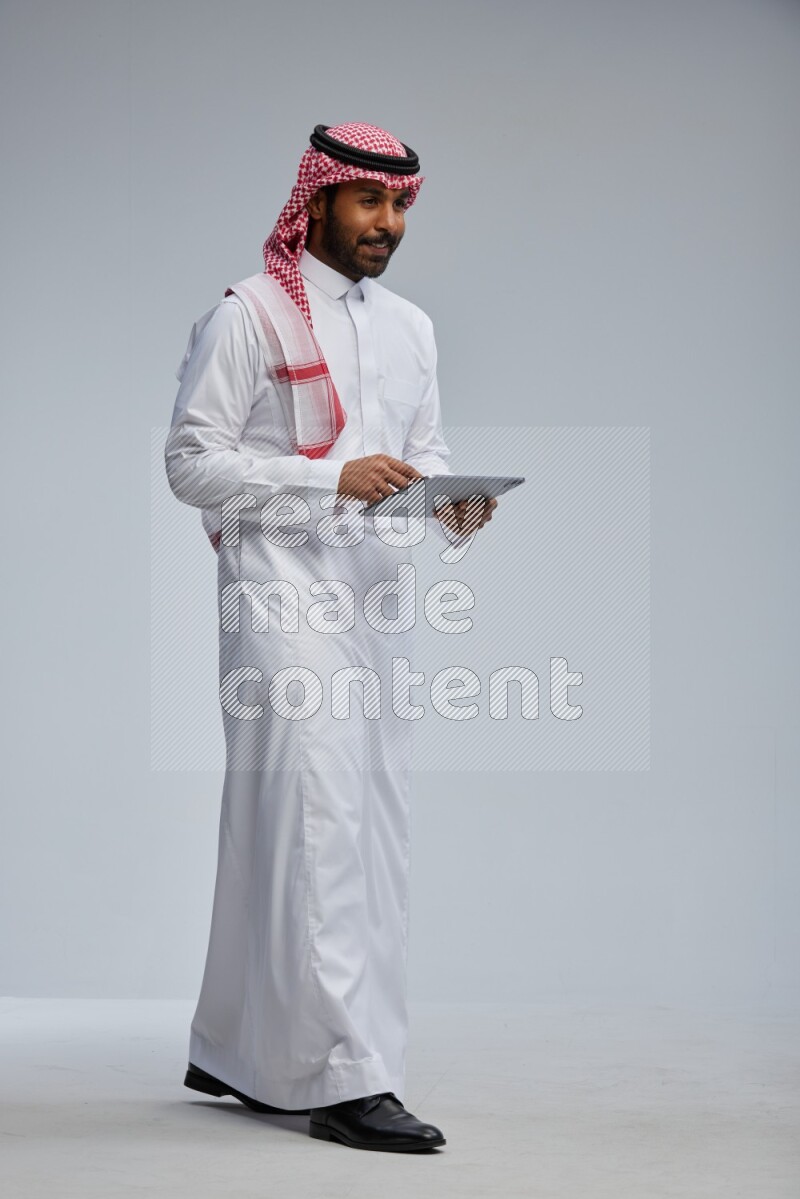 Saudi man Wearing Thob and shomag standing working on tablet on Gray background