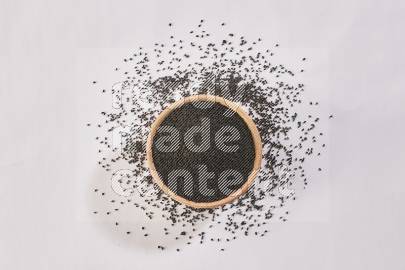 A wooden bowl full of black seeds and more seeds spread on a white flooring