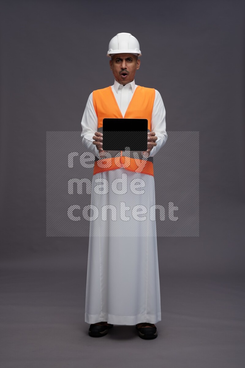 Saudi man wearing thob with engineer vest standing showing tablet to camera on gray background