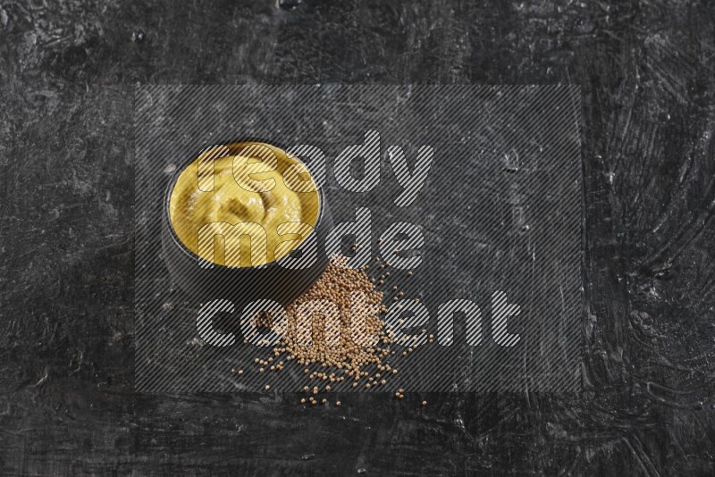 A black pottery bowl full of mustard paste with mustard seeds underneath on textured black flooring