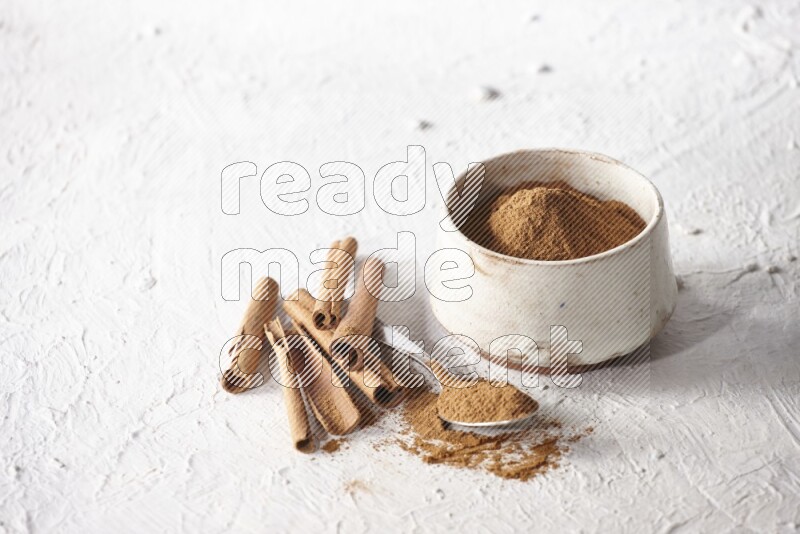Ceramic beige bowl full of cinnamon powder and a metal spoon with cinnamon sticks next of it on a textured white background