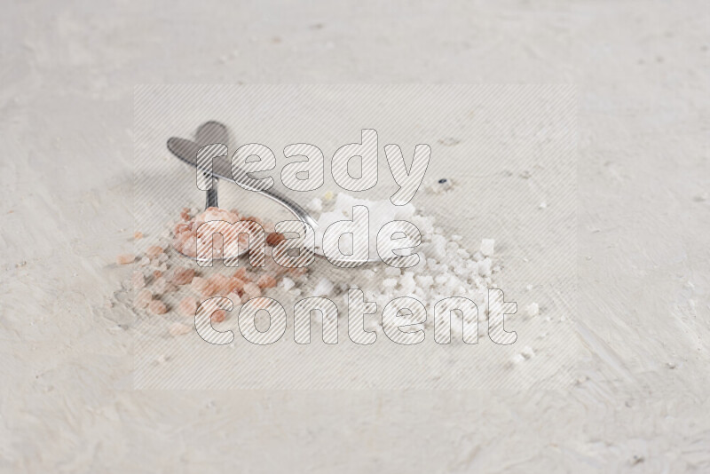 2 metal spoons filled with white salt and pink himalayan salt on white background