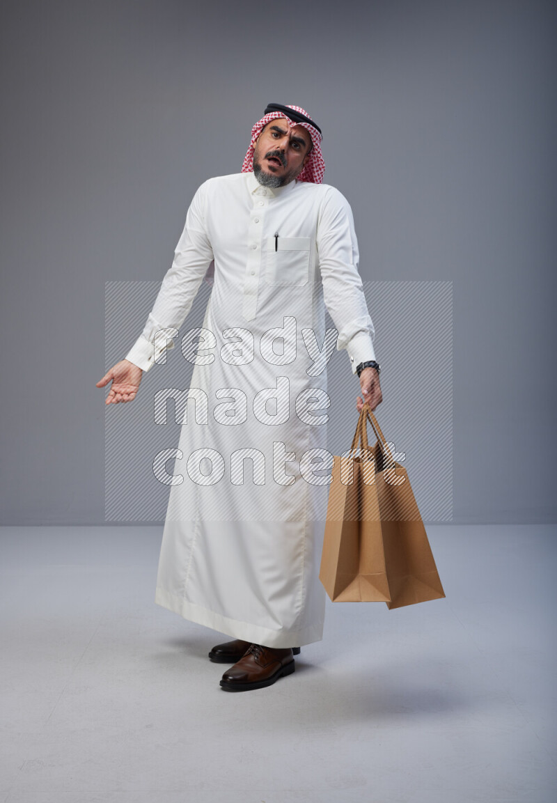 Saudi man Wearing Thob and red Shomag standing holding shopping bag on Gray background